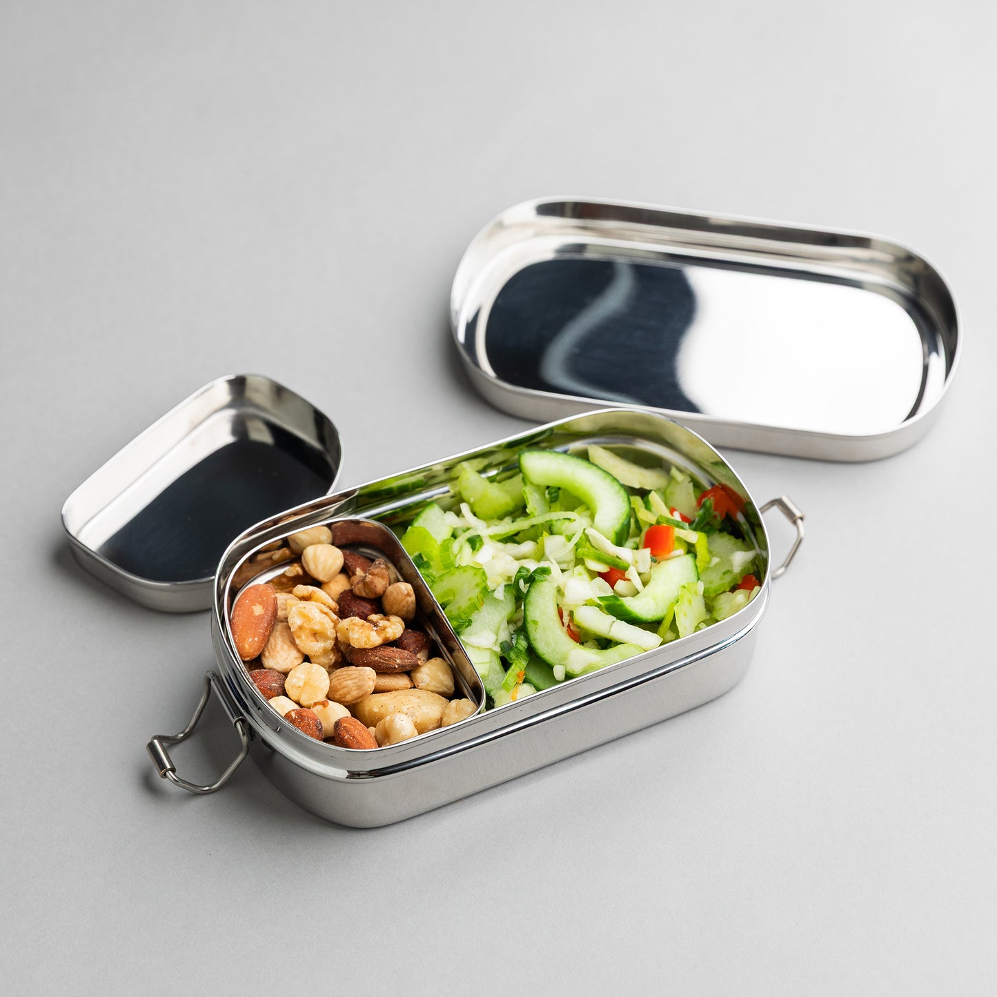 RVS broodtrommel met vakjes gevuld met noten en salade, op een grijze ondergrond