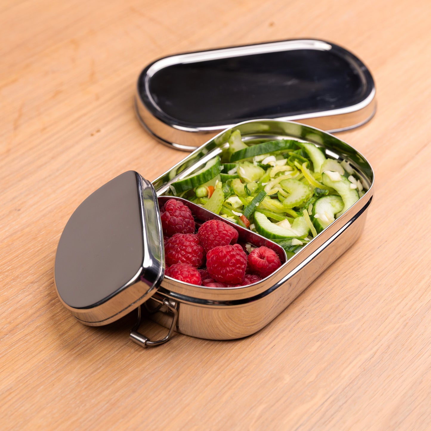 RVS lunchbox met vakjes gevuld met framboos en salade op een schooltafel