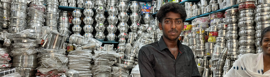 Man in een winkel in india met verschillende RVS producten uit india. Waaronder potten RVS borden, RVS bekers