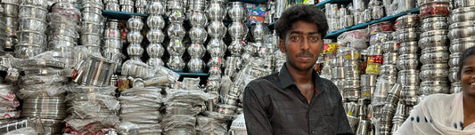 Man in een winkel in india met verschillende RVS producten uit india. Waaronder potten RVS borden, RVS bekers