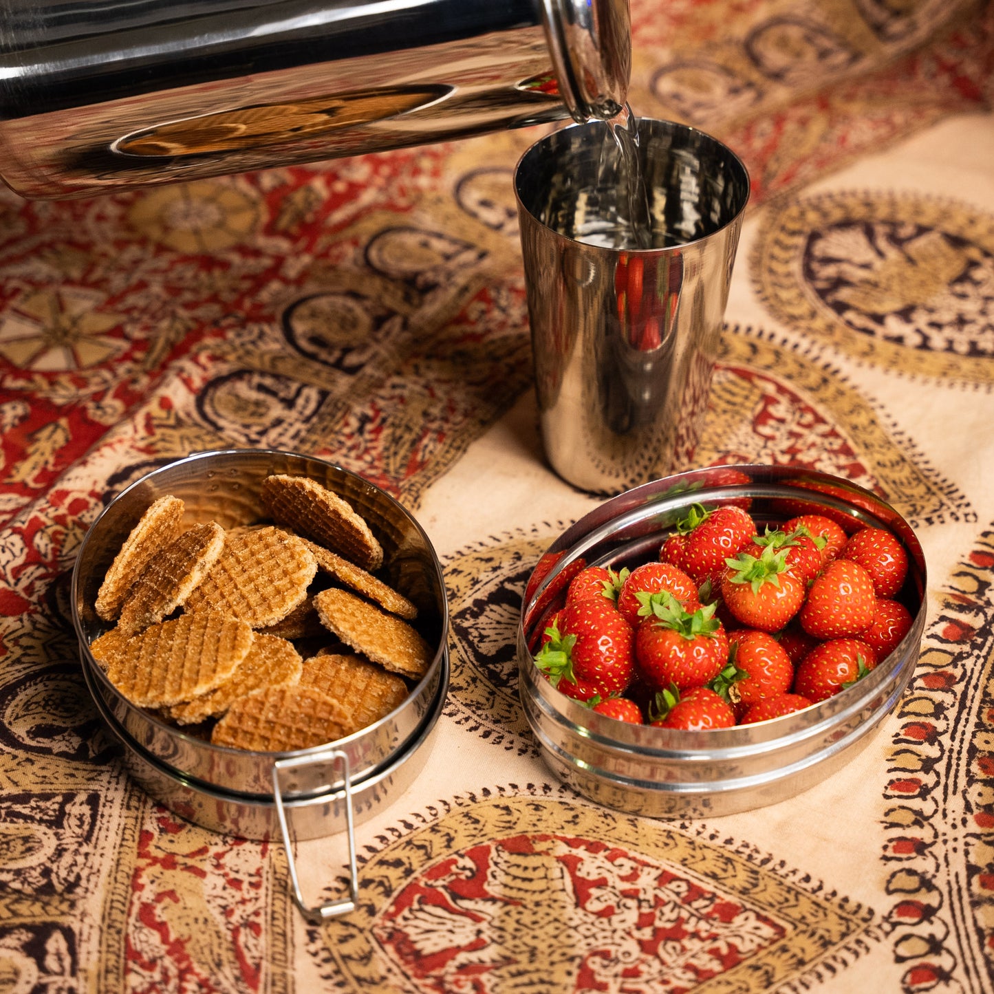 rvs lunchbox met vakjes voor kinderen gevuld met aardbeien en koekjes. op een indiase doek. met waterkan en glas