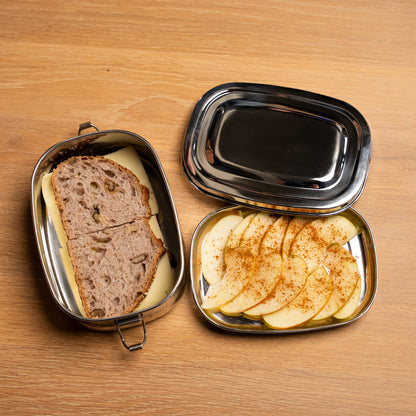 rvs lunchbox en broodtrommel gevuld met een boterham en appeltjes. Op een  tafel van hout. deze broodtrommel is voor kinderen en volwassenen