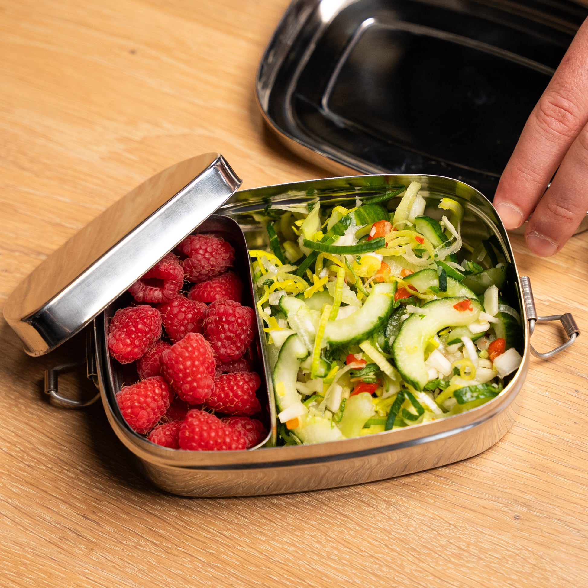 rvs Lunchbox voor kinderen gevuld met sla. in de lunchbox vakjes en een fruitbakje. rechts in beeld 2 vingers