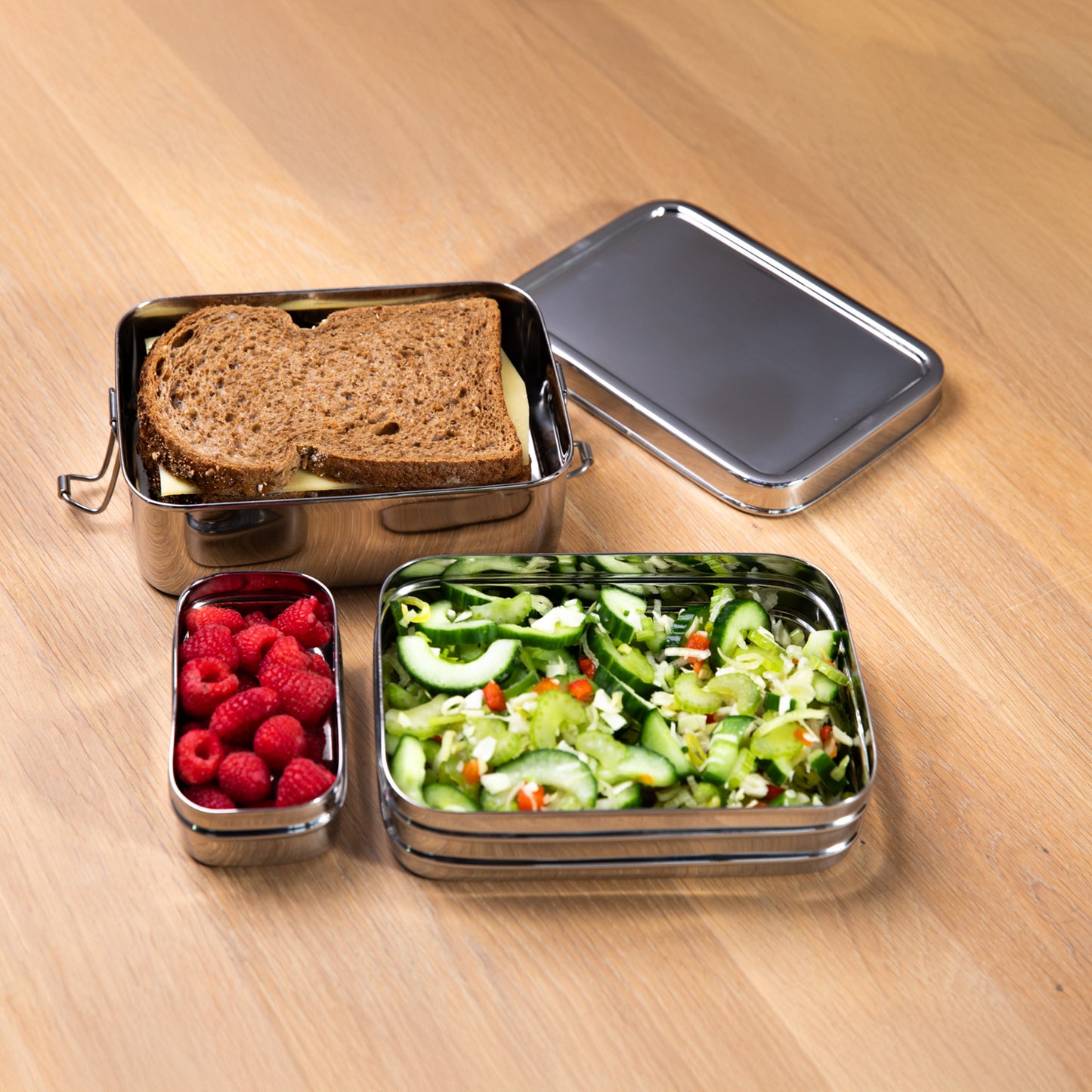 RVS lunchbox met een boterham, sla en frambozen op een houten tafel