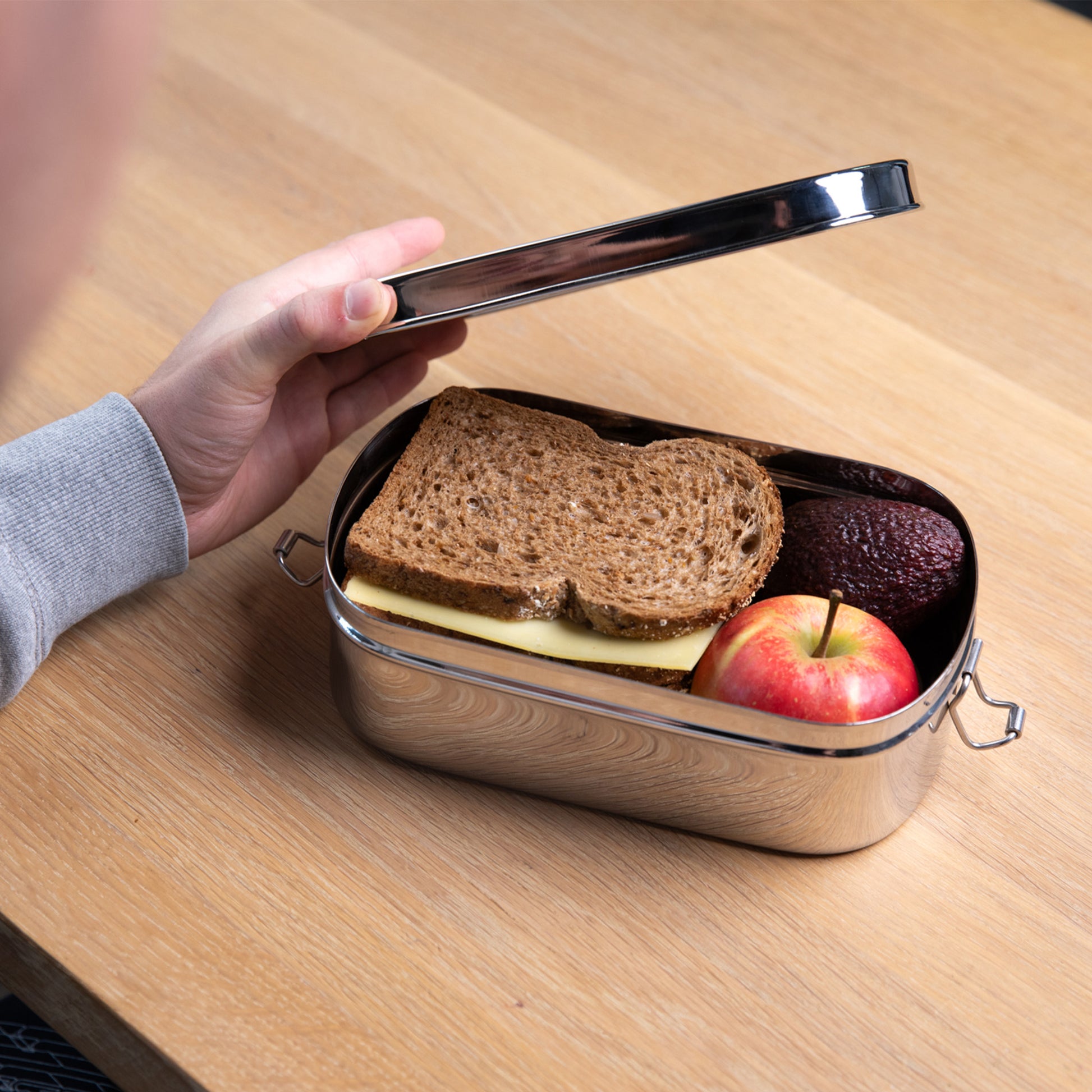 RVS lunchbox die word geopend door een hand. in de lunchbox zit een boterham en een appel